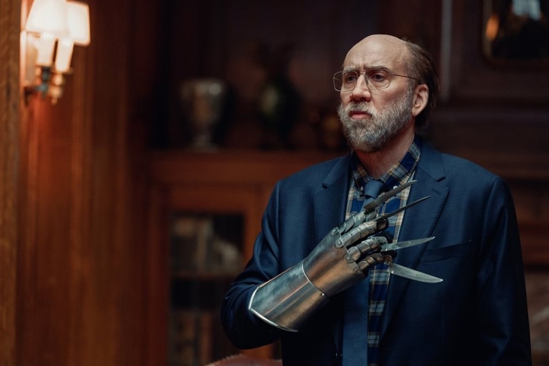 Actor Nicolas Cage, in character, stands solemnly, holding Freddie-Krueger style gloved knife hands over his chest. 
