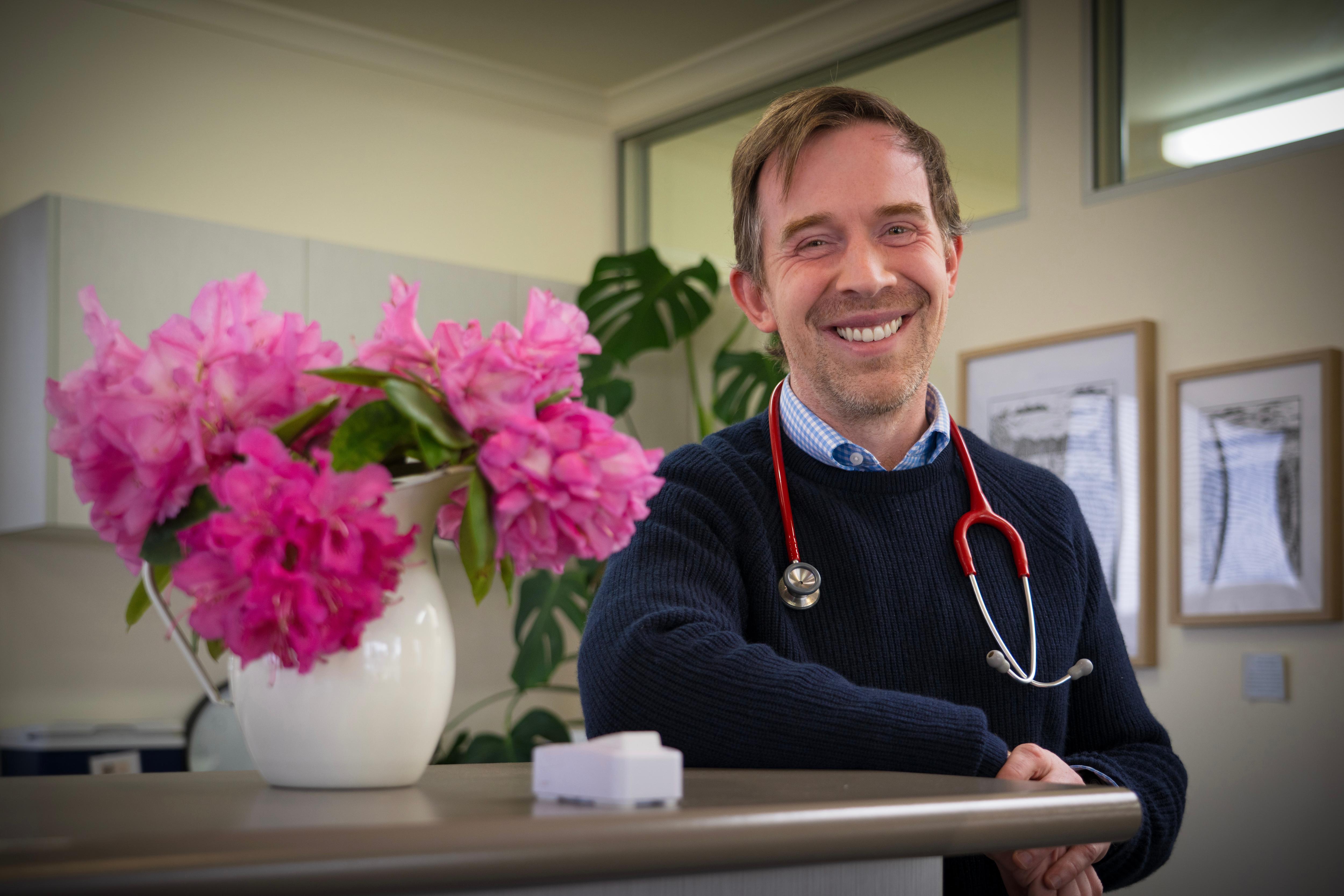 A middle aged white man with a stethoscope around his neck, smiling and leaning on a counter