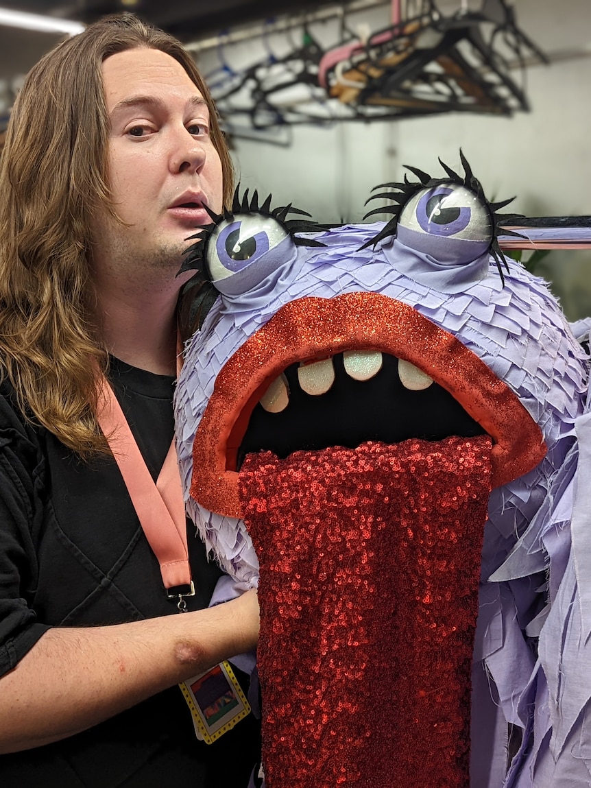 Blake Anderson poses next the head of a purple monster costume with red lips