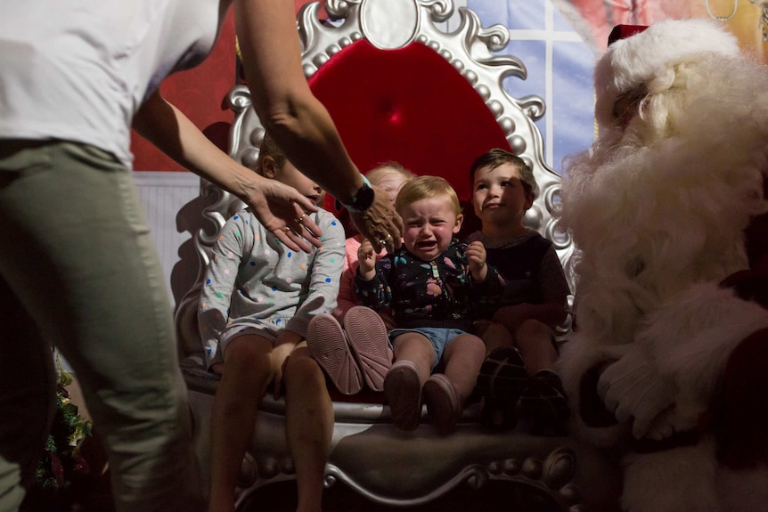 A baby cries on Santa's throne as his mother reaches for him.