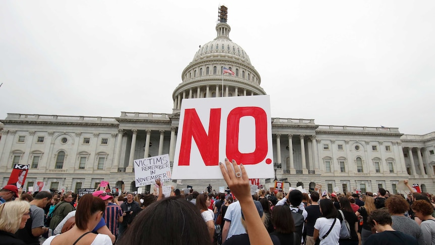 A woman standing in a large crowd holds a sign that says "No." in front of the US Capitol building