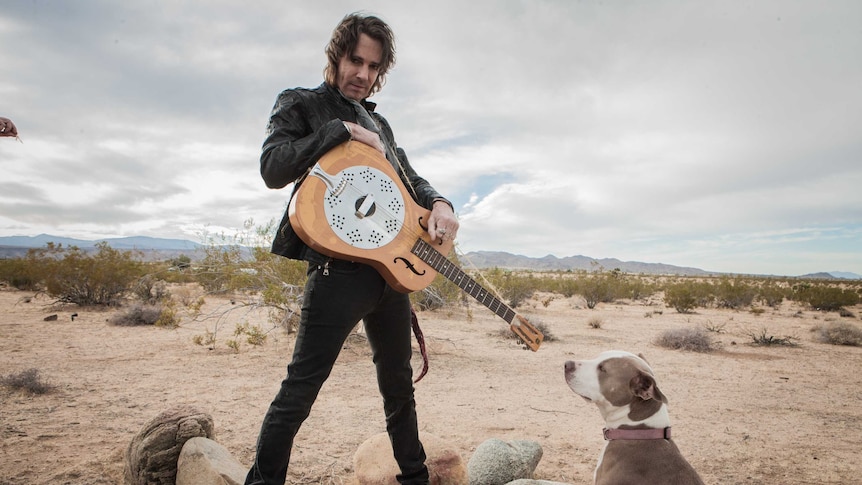 Rick Springfield shot from below holding a guitar in a desert with a dog sitting in the bottom right of the frame.