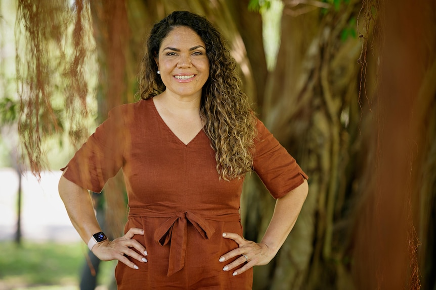 A woman looks at the camera with a serious expression and hands on her hips. She wears a ochre dress.