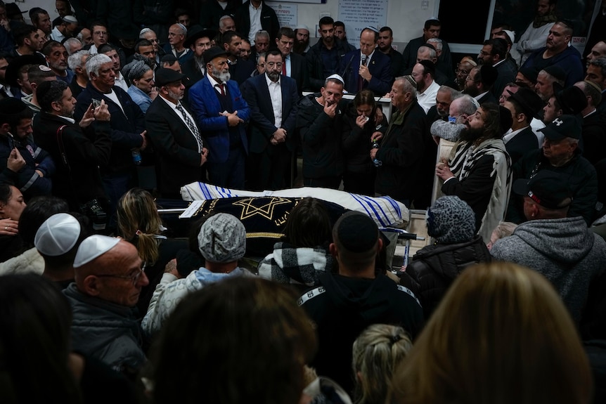 A large crowd of mourners gather around a coffin upon which an Israeli flag has been draped.