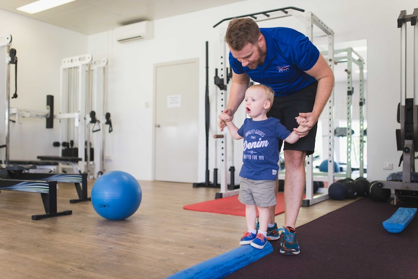 A man holds the hands of a toddler to support him as he walks across a beam.