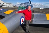 An older man in a red jumper leans, smiling, on a vintage propeller aeroplane on the tarmac.