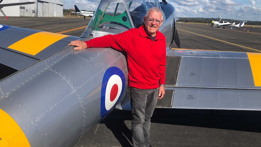 An older man in a red jumper leans, smiling, on a vintage propeller aeroplane on the tarmac.