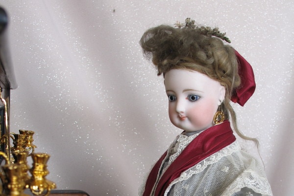 An antique doll dressed in a lacy robe sits at a piano.