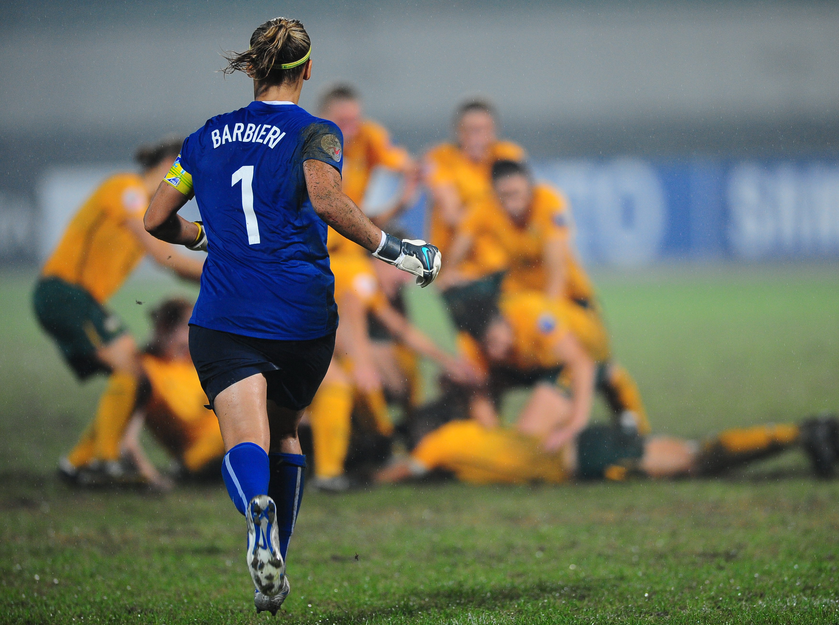 Um goleiro de azul corre em direção a um grupo de jogadores comemorando em verde e dourado em um campo de futebol enlameado
