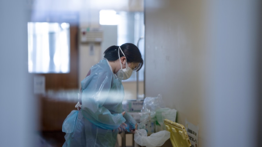 A nurse standing in a hospital hallway pulls off her shower cap while dressed in full PPE 