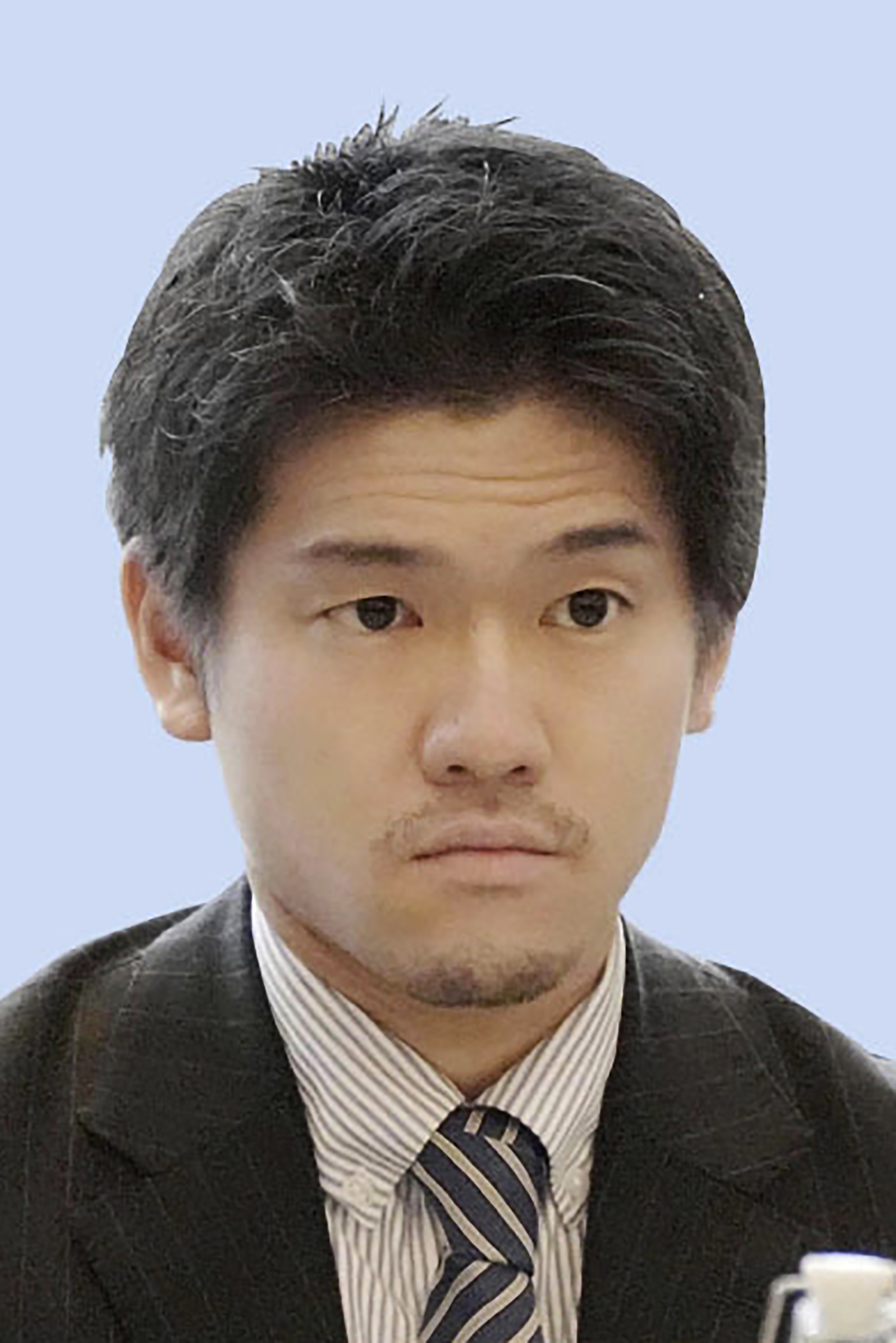 A headshot of a young Japanese man in a suit and tie looking serious against a blue background.