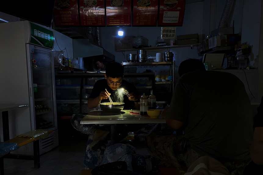 Un hombre come un plato de fideos en una habitación oscura, con solo la luz de su teléfono para iluminar lo que hay en el plato.