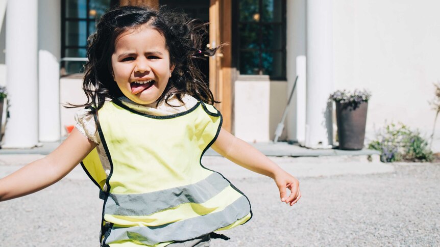 A young girl in a reflective, hi-vis vest plays outside at a childcare centre. She has dark hair and has her tongue stuck out.