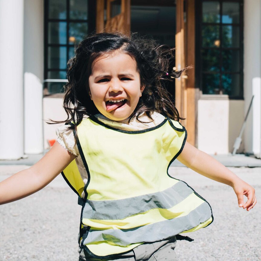 A young girl in a reflective, hi-vis vest plays outside at a childcare centre. She has dark hair and has her tongue stuck out.