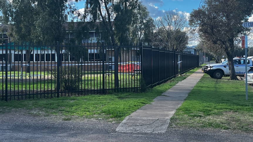 Police tape is stretched across a schoolyard and a police car is parked at the gate.