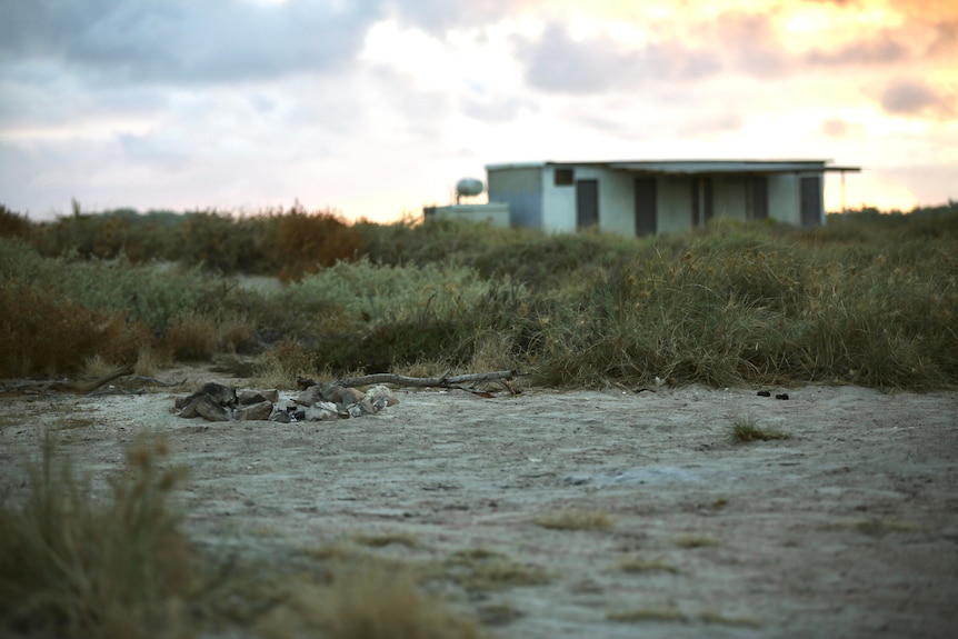 An old campfire on sandy ground in front of beach scrub at dusk, with a tin shack in the background.