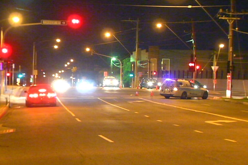 Police cars block of a wide street in Albion as traffic lights shine at night, with orange cones placed on the road.