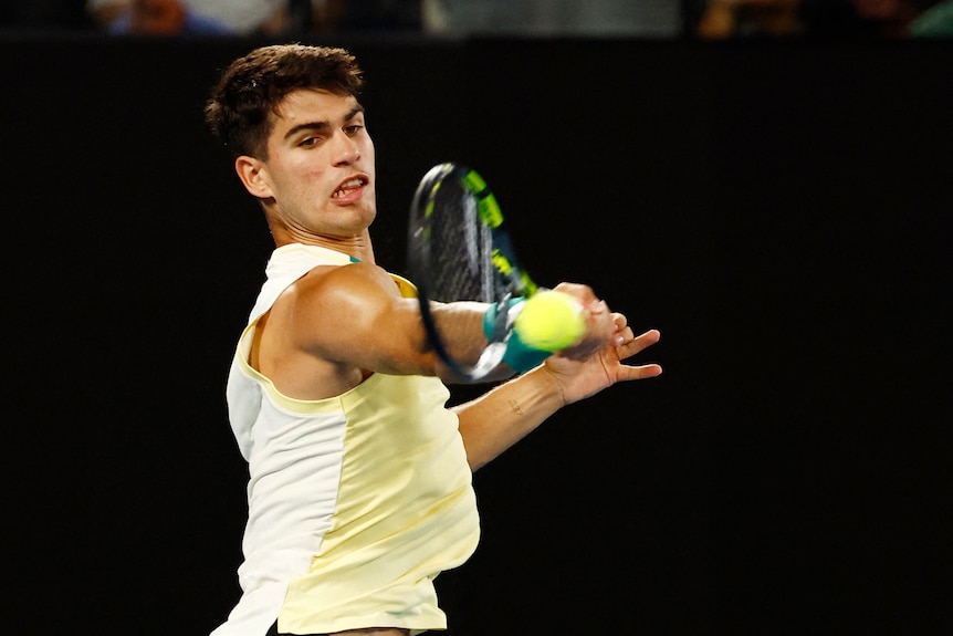 A male tennis player in a yellow singlet plays a forehand, using all of his energy