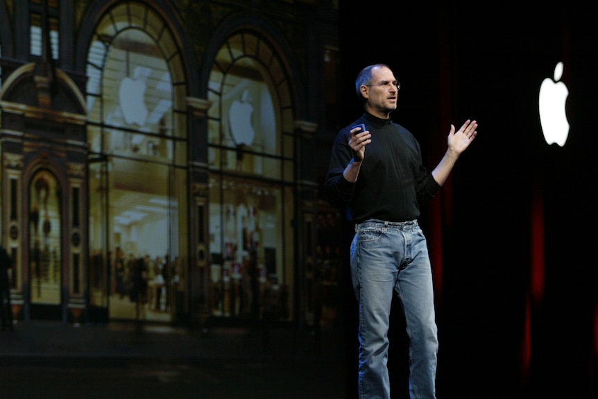 apple ceo steve jobs stands on a stage speaking and holding a small remote. an apple logo can be seen lit up behind him