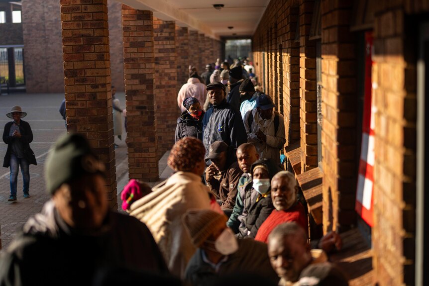 A long line of people stand in an outdoor corridor in the early morning sun
