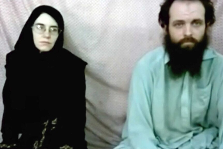 A still image of a woman and man sitting in front of a sheet.