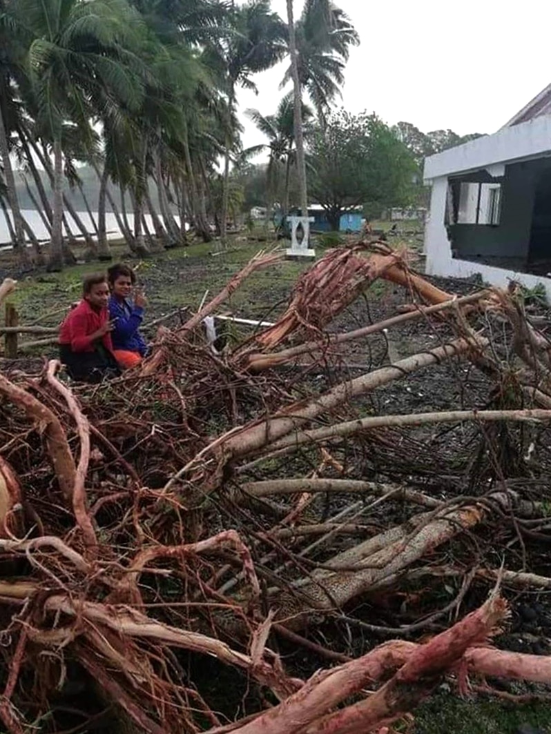 You view mangled tree debris in the foreground while two women make peace signs to the camera as they sit amongst it.