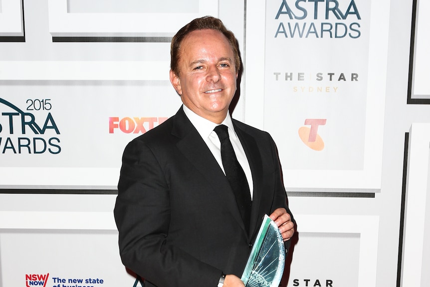 A man in a black suit holds a crystal trophy on a red carpet