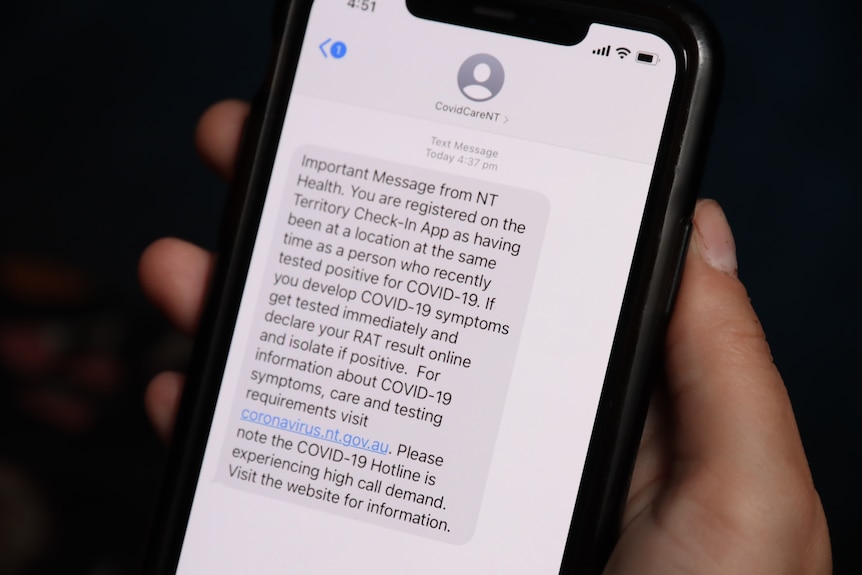 A hand holding a phone displaying a COVID-19 contact tracing text message from the NT government.