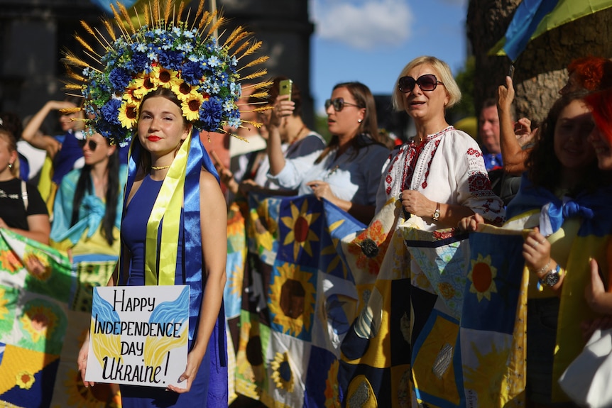 A woman in a yellow and blue headdress holds a sign reading "happy independence day Ukraine" 