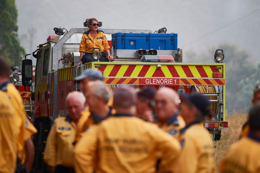 Female firefighter sitting on top of a fire truck, behind out of focus firefighters standing in front