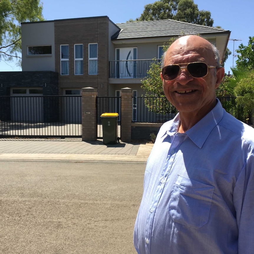 An older bald man wearing sunglasses standing in front of a modern two-storey house