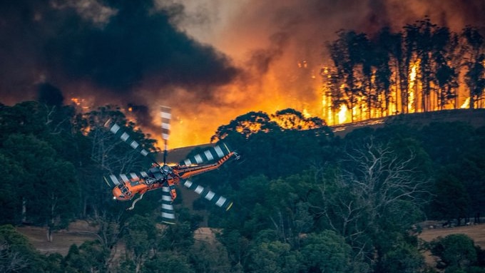 An orange helicopter flies over burning bushland.
