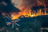 An orange helicopter flies over burning bushland.