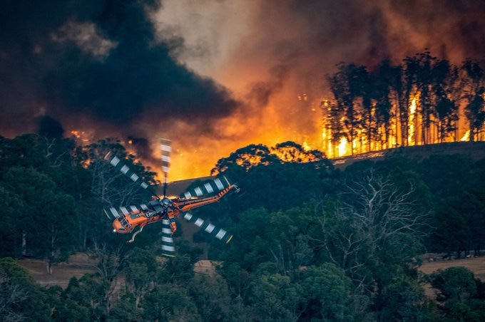 An orange helicopter flies over burning bushland.