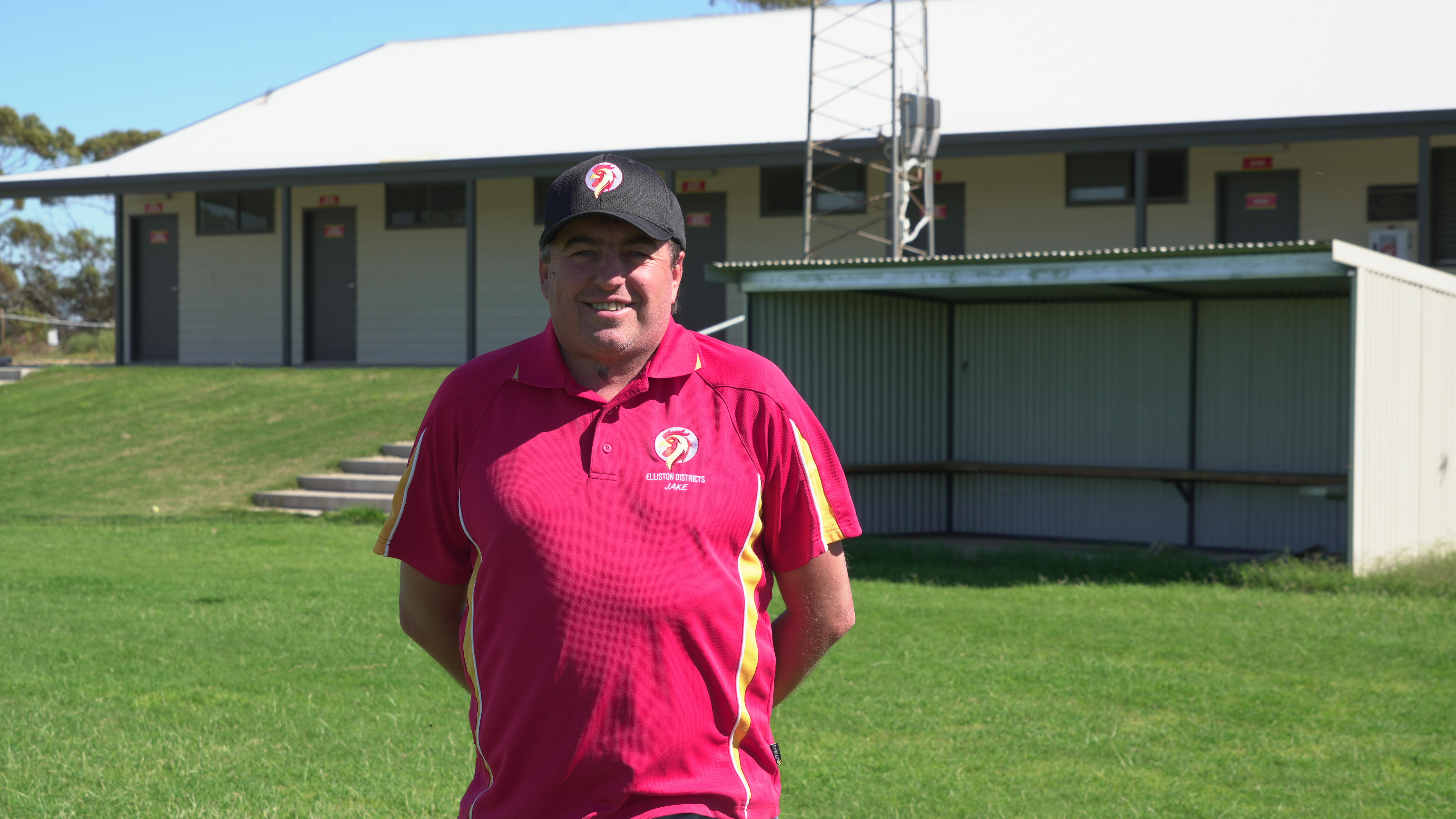 Homem de camisa vermelha em pé sobre um oval verde em frente ao prédio do clube esportivo 