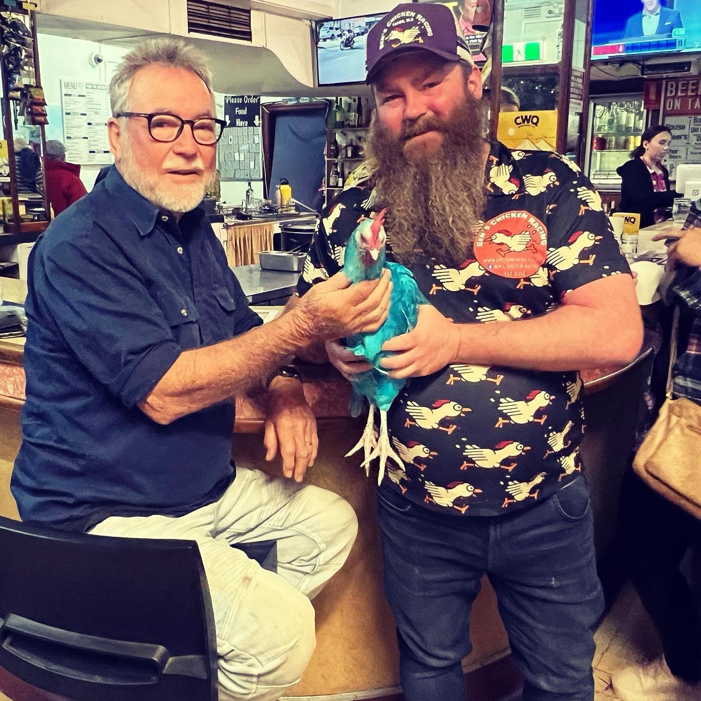 Two men sitting at a bar, one on left with a beard and holding a chicken