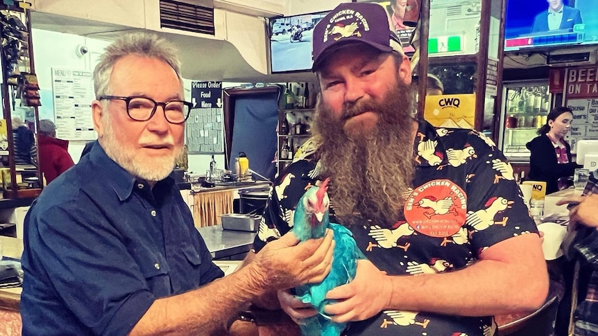 Two men sitting at a bar, one on left with a beard and holding a chicken