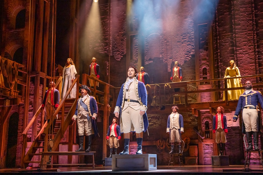  Spotlit stage with men in 18th century military uniform, standing on chairs and boxes and tables, singing.