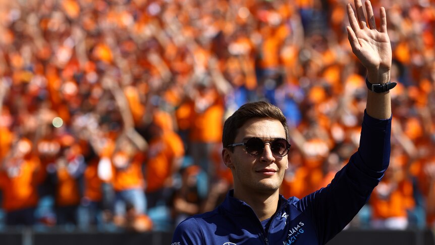 George Russell, wearing sunglasses, waves to a crowd of people wearing orange