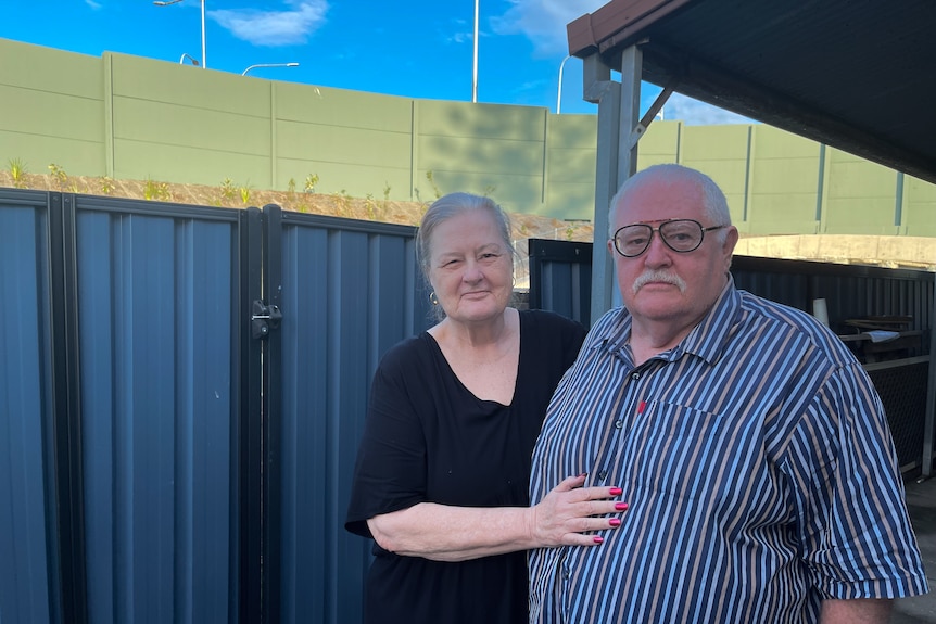 An elderly couple standing in front of a blue steel fence with green highway sound barriers in the background.