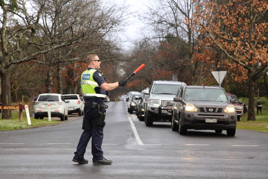 A policeman in a yellow vest waves an orange baton to redirect traffic.