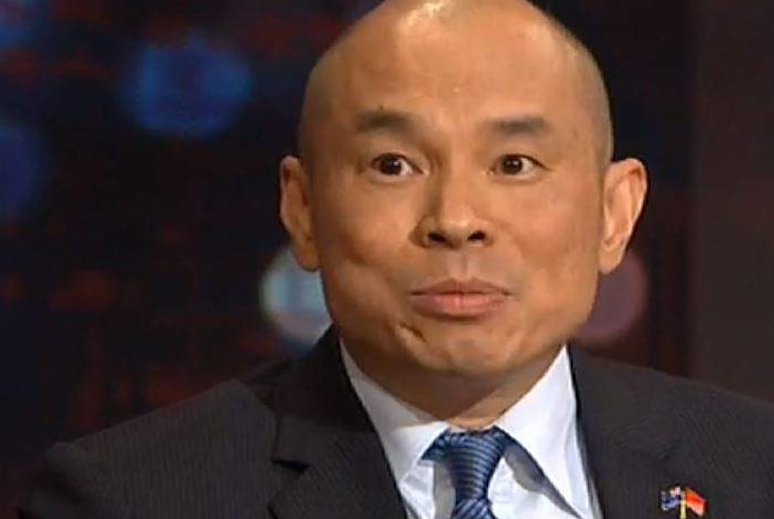 A bald man in a suit and tie with a badge on his lapel looks forward.