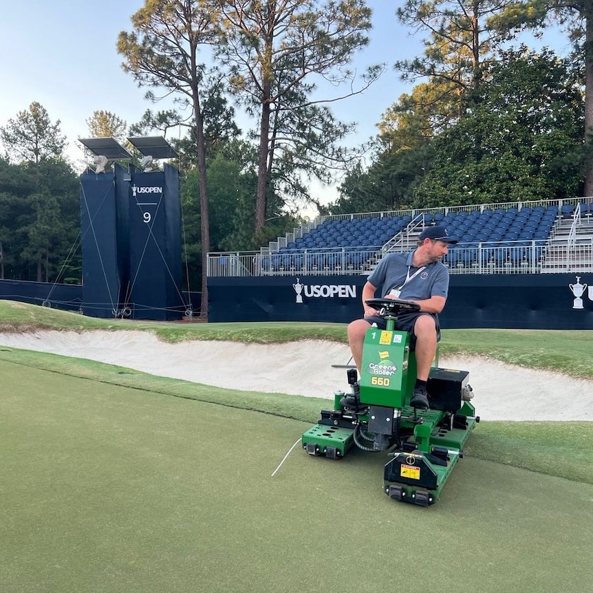 Man working hard on ride-on mower with US Open stands in the background