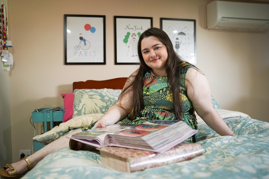 Grace wears a floral patterned dress, sitting on her bed smiling with a photo album.