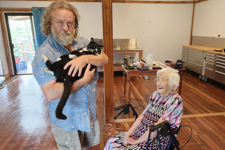 An elderly woman in a wheelchair smiling next to a man holding a black cat.