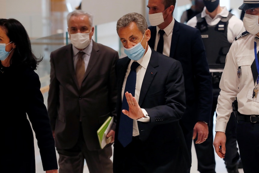 Nicolas Sarkozy wearing a mask walks down a corridor with several other people including a court officer.