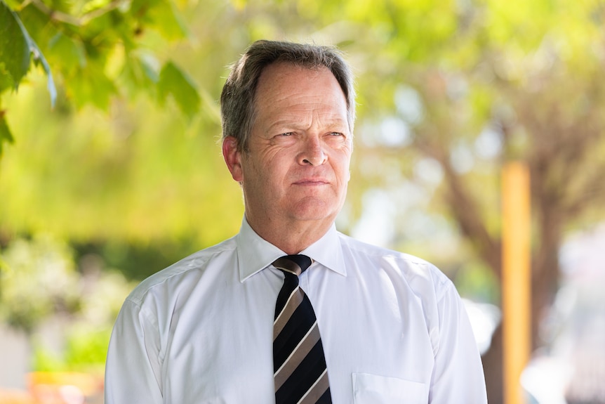 A man in a white shirt with a black and gold tie stands outside looking into the distance.