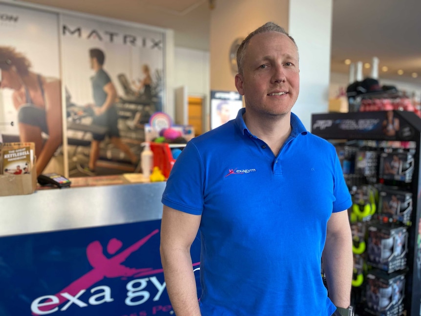 A man in a blue polo shirt standing in a shop with home gym equipment looks into the camera
