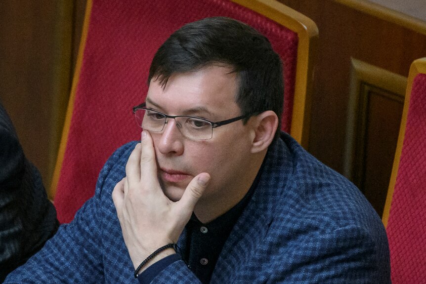 A middle aged man with dark hair and glasses touches his face with his left hand and looks thoughtfully on a red velvet seat