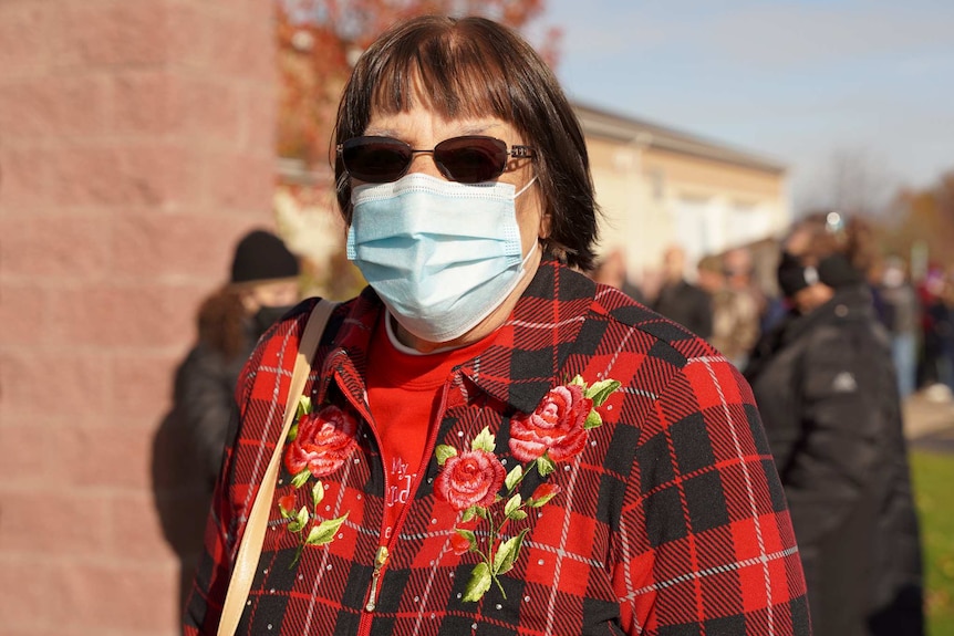 a woman in a red checked jacket with roses on it, wearing sunglasses and a blue face mask stands outside and looks toward camera
